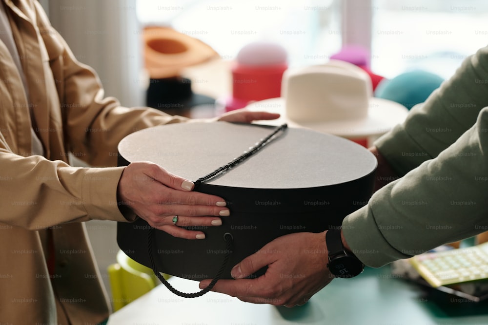 Close-up of young female clerk of craft shop passing black round box with packed handmade product to male client in front of camera