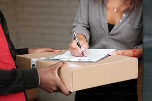 Main d’une jeune femme avec un stylo signant un document sur la réception du colis sur une boîte en carton emballée avec commande livrée par courrier