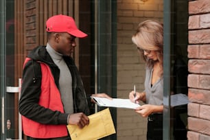 Giovane uomo nero in uniforme che tiene la busta con la lettera mentre la donna d'affari con la penna firma il documento di consegna tenuto dal corriere