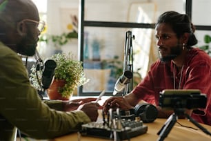 Young intercultural man looking at African American guy during conversation by desk in studio while both talking in microphones