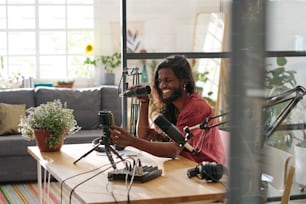 Smiling guy in casualwear switching smartphone while sitting by desk in front of microphone and going to make livestream or record