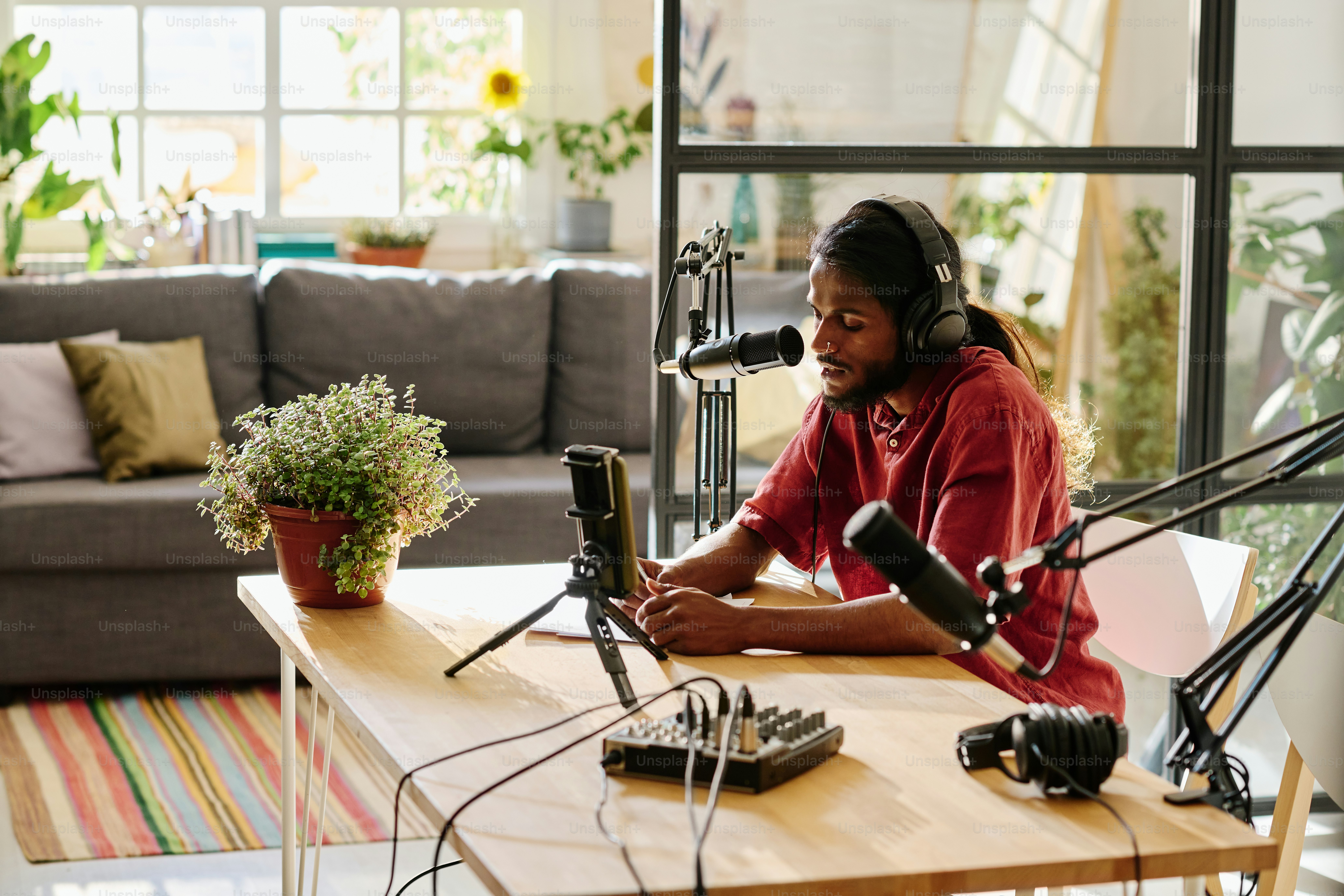 Young man in headphones sitting by desk and reading points of discussion while creating new audio file for online audience