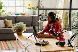 Young man in headphones sitting by desk and reading points of discussion while creating new audio file for online audience