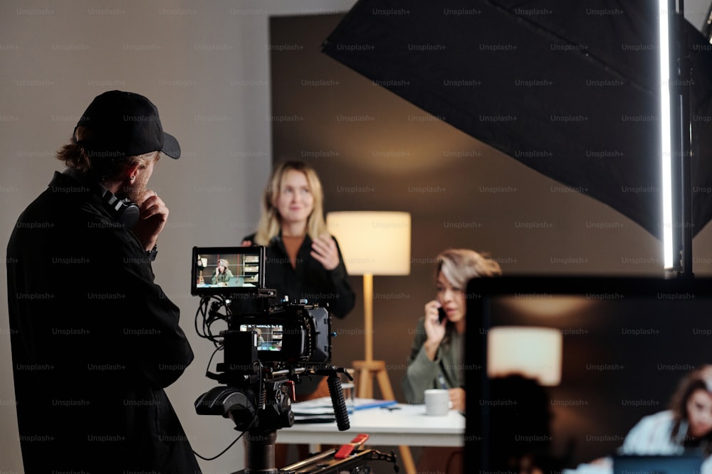 Back view of young videographer standing in front of steadicam during shooting of commercial with female model in studio