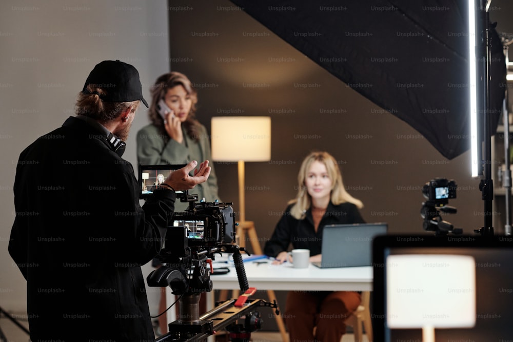 Vista de Raer de jovem cinegrafista em traje casual preto dando conselhos a modelos femininas que participam da filmagem de novo comercial