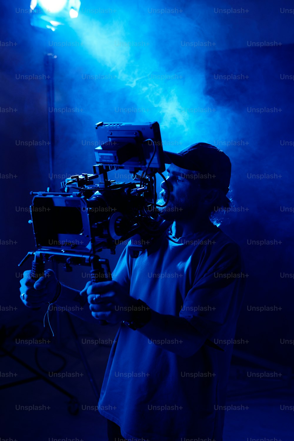 Giovane cameraman serio con steadicam in piedi contro i riflettori in studio illuminato da luce blu e pieno di fumo durante le riprese video