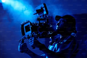 Jovem cinegrafista em casualwear filmando vídeo comercial em sala escura ou estúdio cheio de fumaça iluminada pela luz azul escura