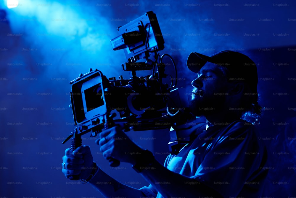 Giovane videografo in abbigliamento casual che gira video commerciali in camera buia o studio pieno di fumo illuminato da luce blu scuro
