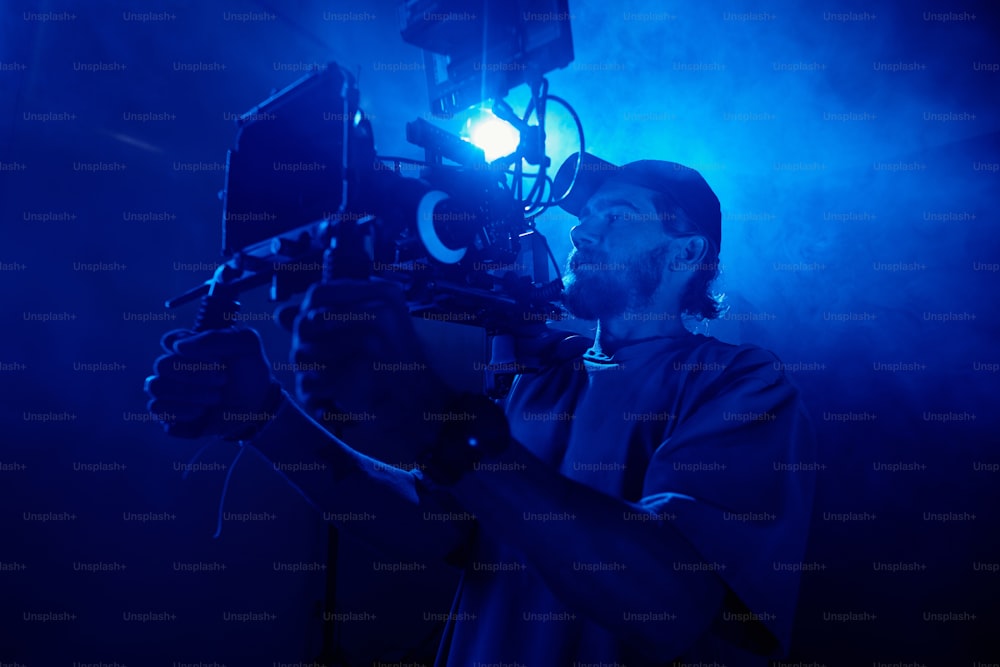 Young man in casualwear holding video camera while shooting commercial in studio lit with blue light and full of smoke