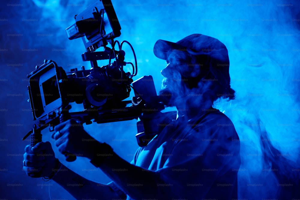 Bearded videographer in baseball cap holding steadicam while standing in smoke and making all the necessary adjustments before shooting