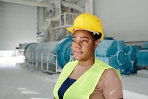 Young serious black woman in uniform and protective helmet standing in front of camera in workshop against machinery equipment