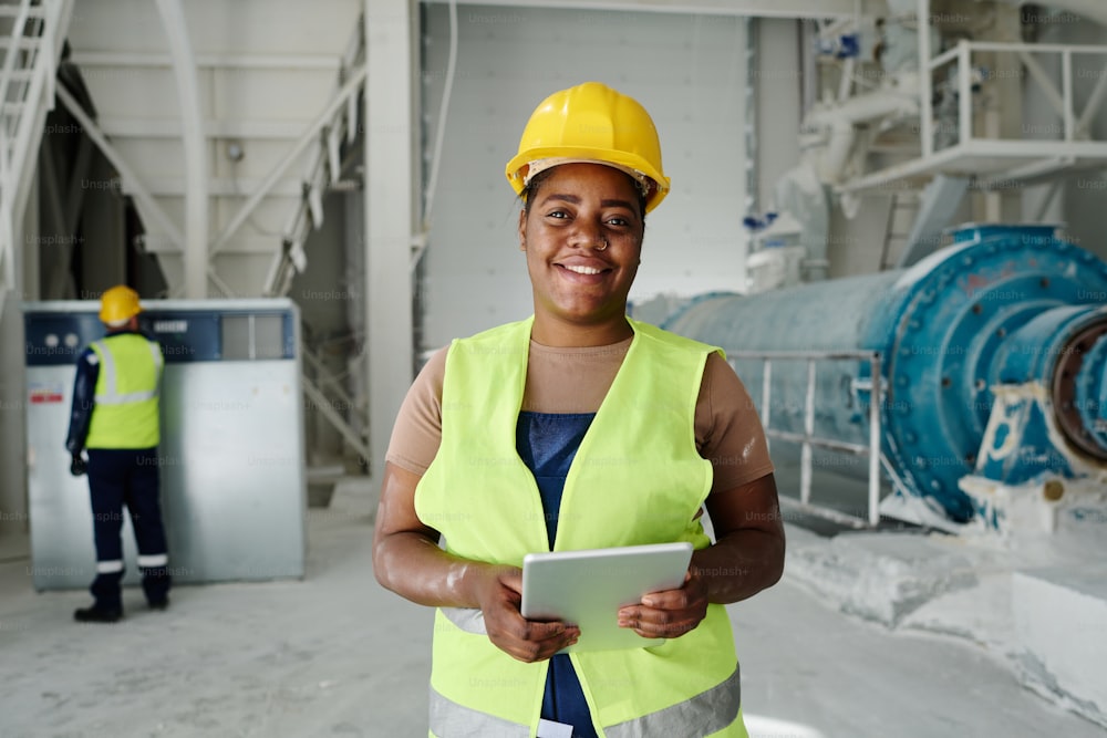 Glückliche junge Afroamerikanerin mit Tablet, die in Produktionsstätte oder Werkstatt vor der Kamera steht und sich vernetzt