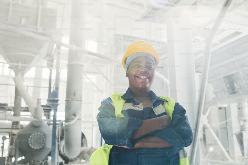 Young successful black female engineer in workwear and protective helmet crossing arms on chest while standing in front of camera