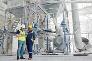 Zwei junge multikulturelle Kollegen in Arbeitskleidung und Schutzhelmen blättern während der Arbeit in einer modernen Fabrik durch eine Online-Anleitung