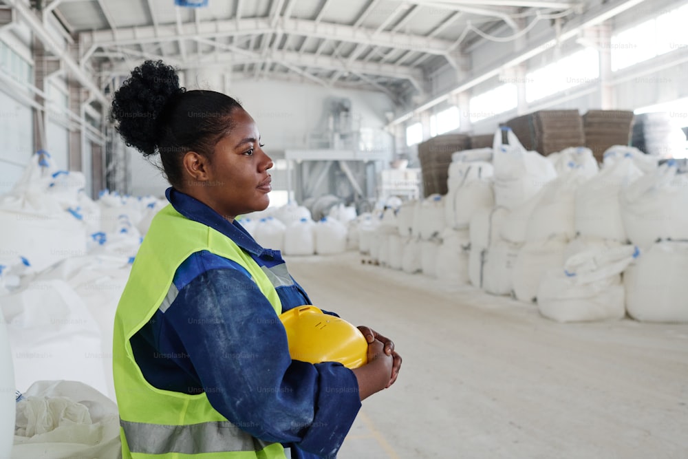 Side view of young black woman in workwear holding yellow safety helmet while standing in front of camera in warehouse