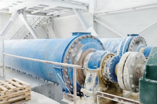 Parte de uma enorme máquina industrial com grande eixo metálico azul durante o processo de produção de mármore em oficina ou planta espaçosa branca