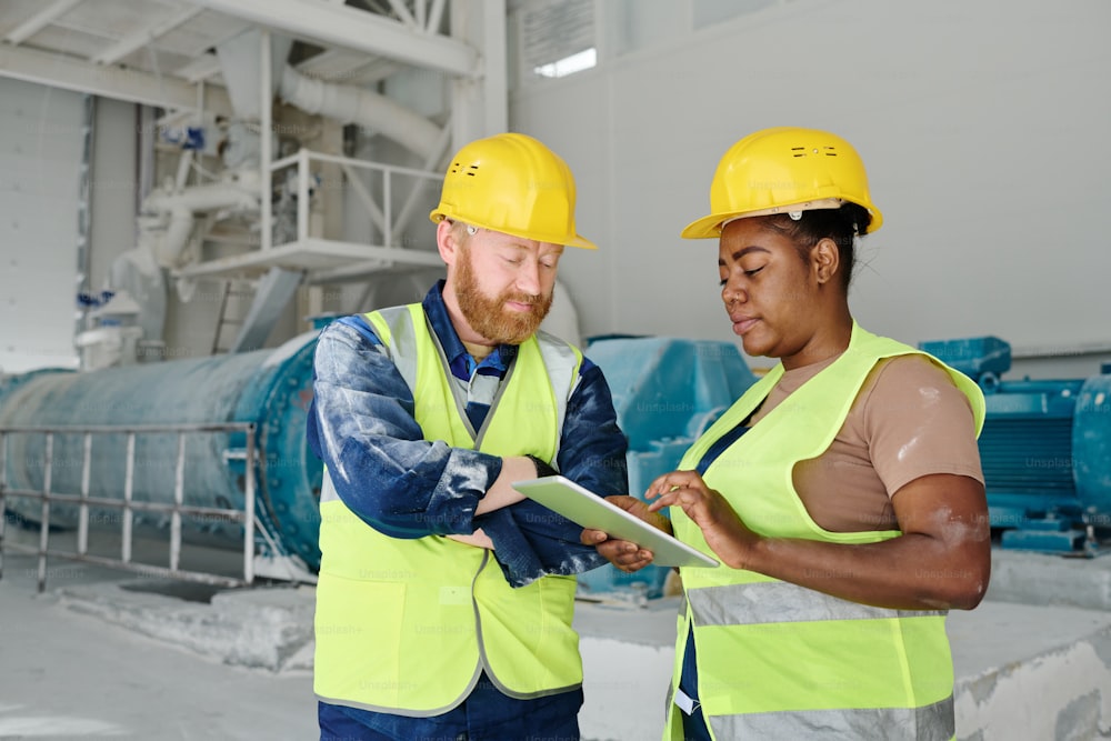 Two intercultural engineers in workwear and safety helmets networking or looking through electronic sketch or construction plan