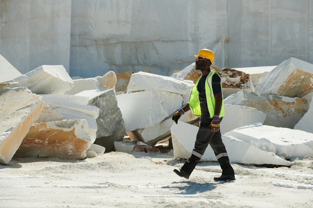 Junger afroamerikanischer männlicher Ingenieur in Arbeitskleidung und Schutzhelm eilt zum Arbeitsplatz, während er sich entlang der dicken Felswand der Fabrik bewegt