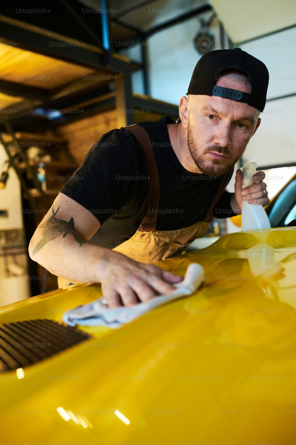 Young mechanic or technician of maintenance service cleaning cover of hood with detergent while bending over front part of car body