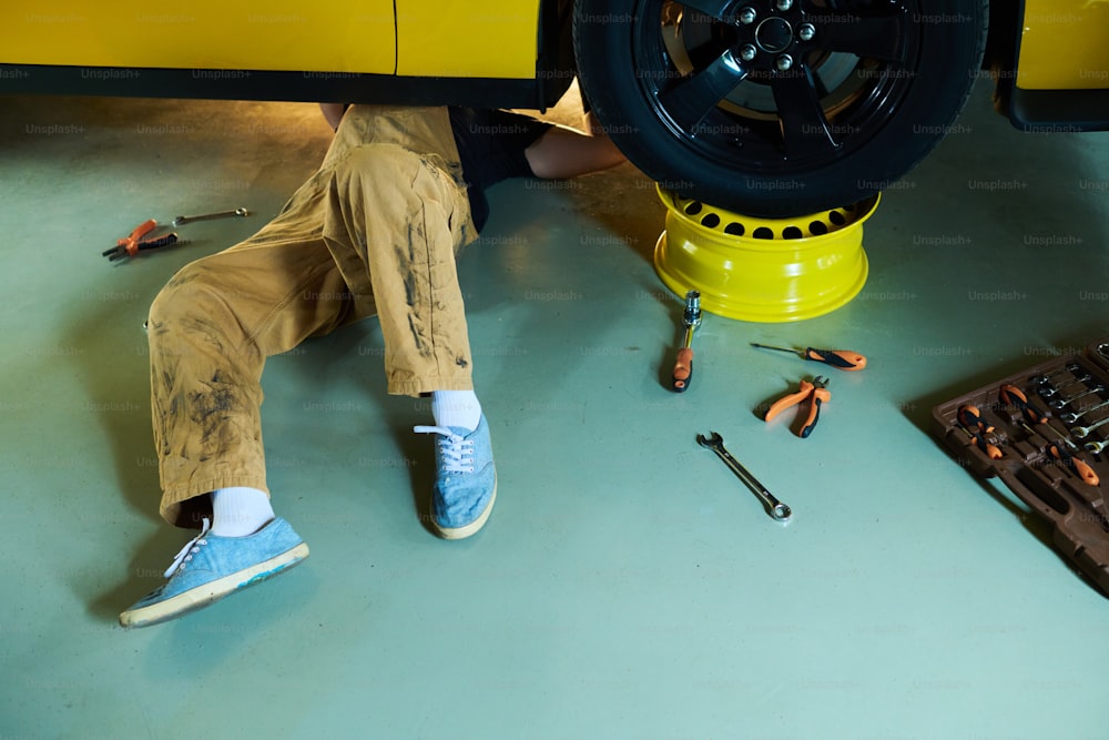 Gambe di giovane riparatore o tecnico in tuta che giacciono sotto l'auto elettrica gialla sul pavimento dell'officina o della garae e la riparano