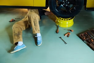 オーバーオールを着た若い修理工や技術者の足が、ワークショップやガラエの床にある黄色い電気自動車の下に横たわって修理している