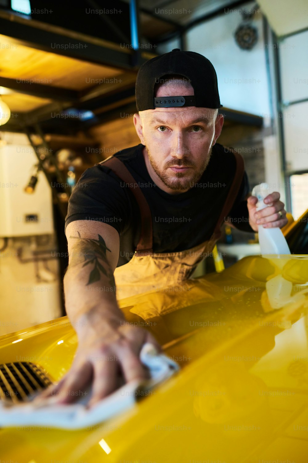 Joven trabajador del servicio de mantenimiento con botella de detergente limpieza cubierta del capó del automóvil amarillo después de los trabajos de reparación