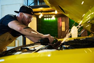 ガレージでの作業中にモーターの詳細を修理するために電気グラインダーを使用しながら、車のエンジンを曲げる若い修理工