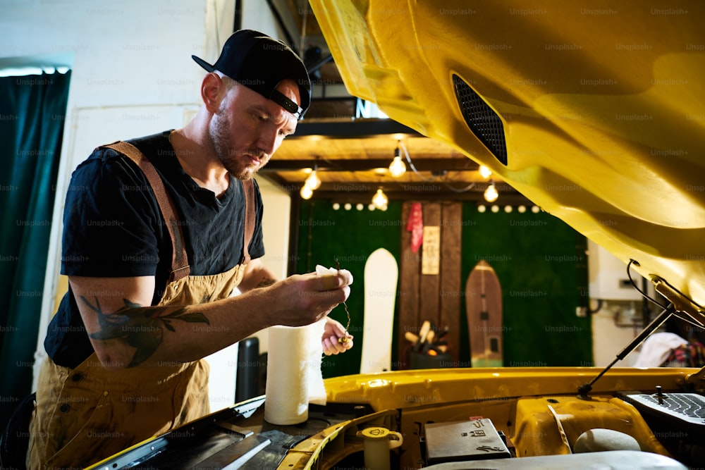 Jeune homme sérieux en tablier et casquette essuyant du fil métallique avec une serviette en papier tout en se tenant près d’une voiture cassée avec capot ouvert dans un garage