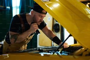 自動車修理工場の若い整備士が、モーターにフラッシュライトを向けながら黄色の自動車のエンジンをチェックする