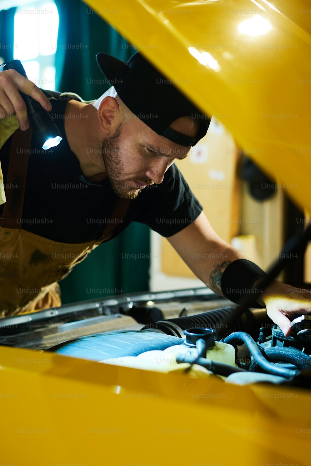 黄色い車の開いたボンネットの上に懐中電灯を曲げ、欠陥をチェックし、ガレージのモーターの部品を固定する真面目な自動車整備士