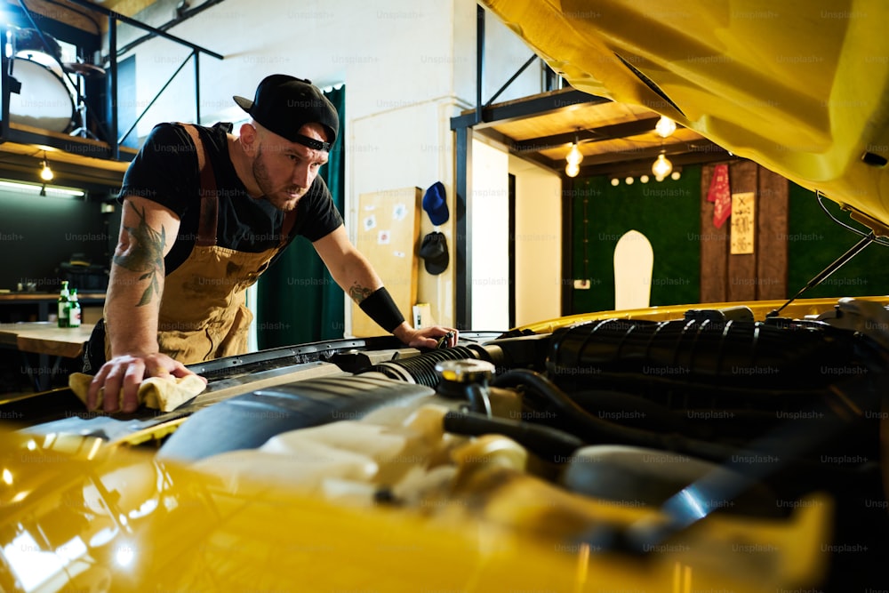Young serious man in apron and baseball cap bending over open hood of car and carrying out checkup of engine in garage or workshop