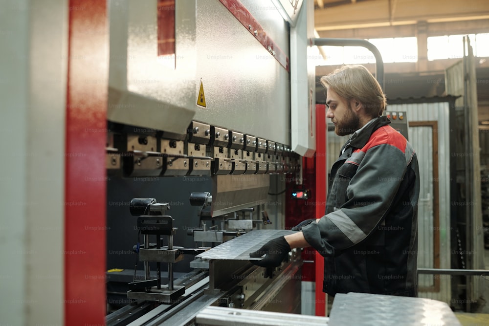 Vista lateral de un joven ingeniero en ropa de trabajo colocando láminas metálicas en parte de una máquina industrial en funcionamiento u otro equipo en la fábrica
