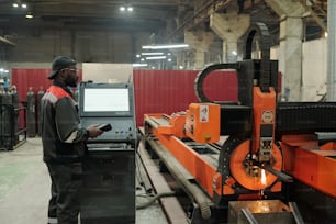 Young male worker of industrial plant standing by control terminal while checking work of production machine in workshop