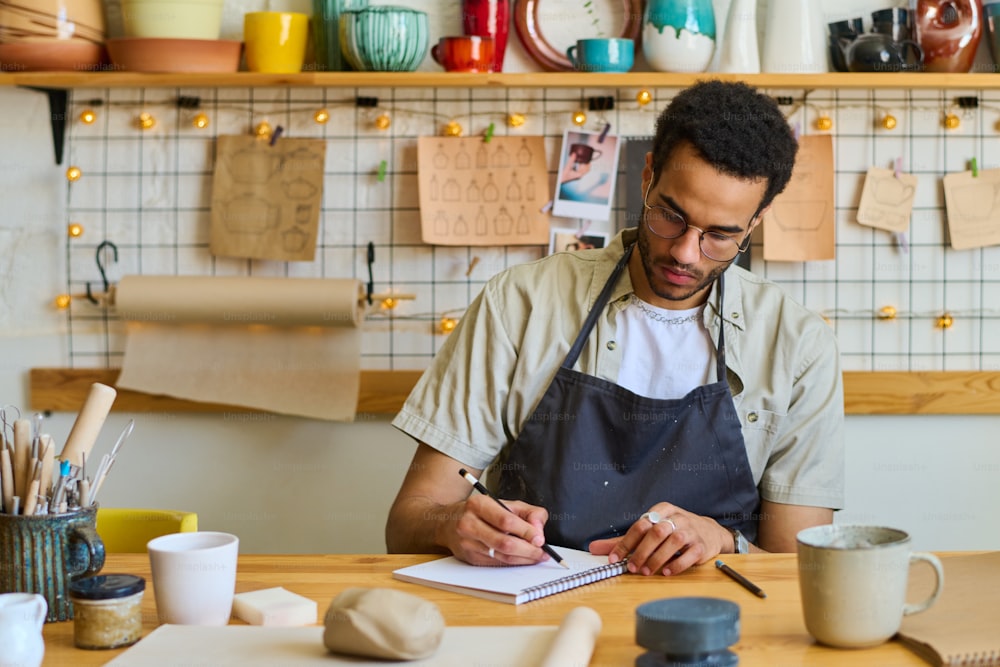 Junger kreativer schwarzer Mann, der Skizzen von neuen handgefertigten Bastelgegenständen in Notizblock zeichnet oder Arbeitsnotizen macht, während er am Tisch im Studio sitzt