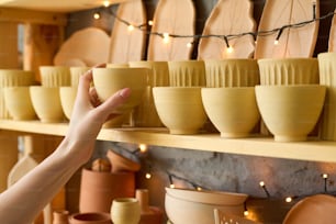 Hand of young female customer taking handmade clay cup or bowl from wooden shelf with other earthenware in small pottery shop