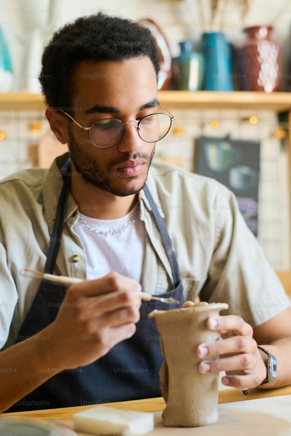 Serious guy in eyeglasses and apron using handtool while creating clay jug or vase for collection of earthenware items for sale