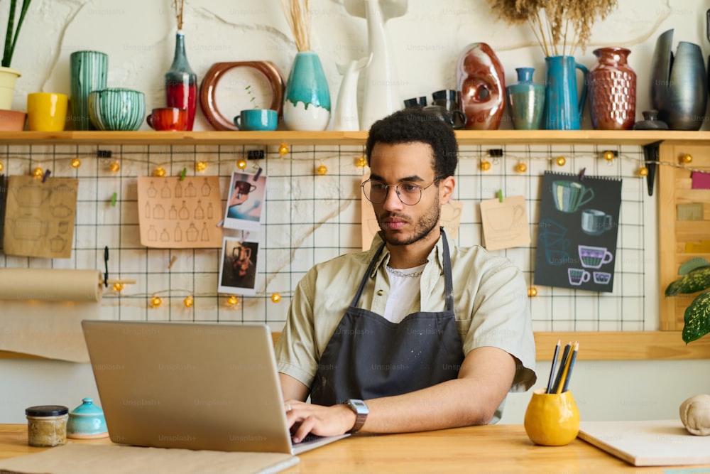 作業服と眼鏡をかけた若い真面目な黒人男性が、職場のそばでノートパソコンの前に座り、クライアントの新しいオンライン注文を探している