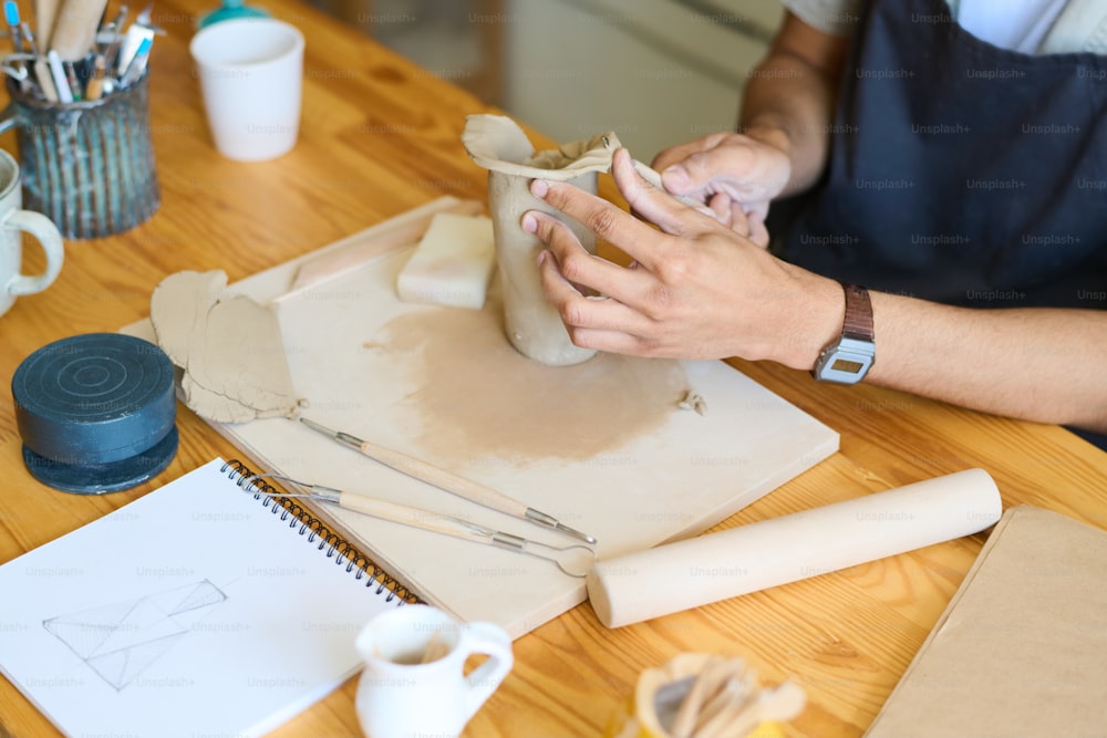 Mãos de oleiro macho jovem formando forma de vaso ou jarro enquanto sentado à mesa e trabalhando sobre novos itens de argila artesanais para venda