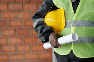 Midsection of foreman in reflective jacket holding protective helmet and blueprint while standing in front of camera against brick wall