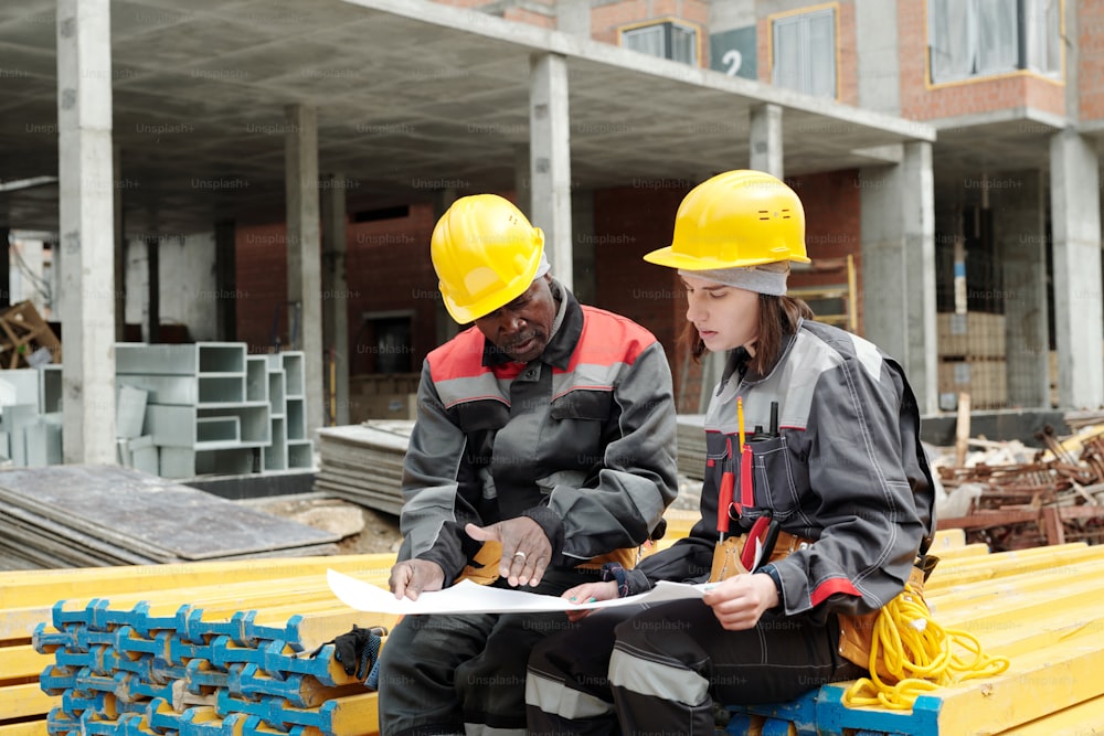 Des constructeurs jeunes et matures en vêtements de travail et casques de sécurité discutent d’un croquis de bâtiment ou d’un plan architectural sur papier lors d’une réunion sur le lieu de travail
