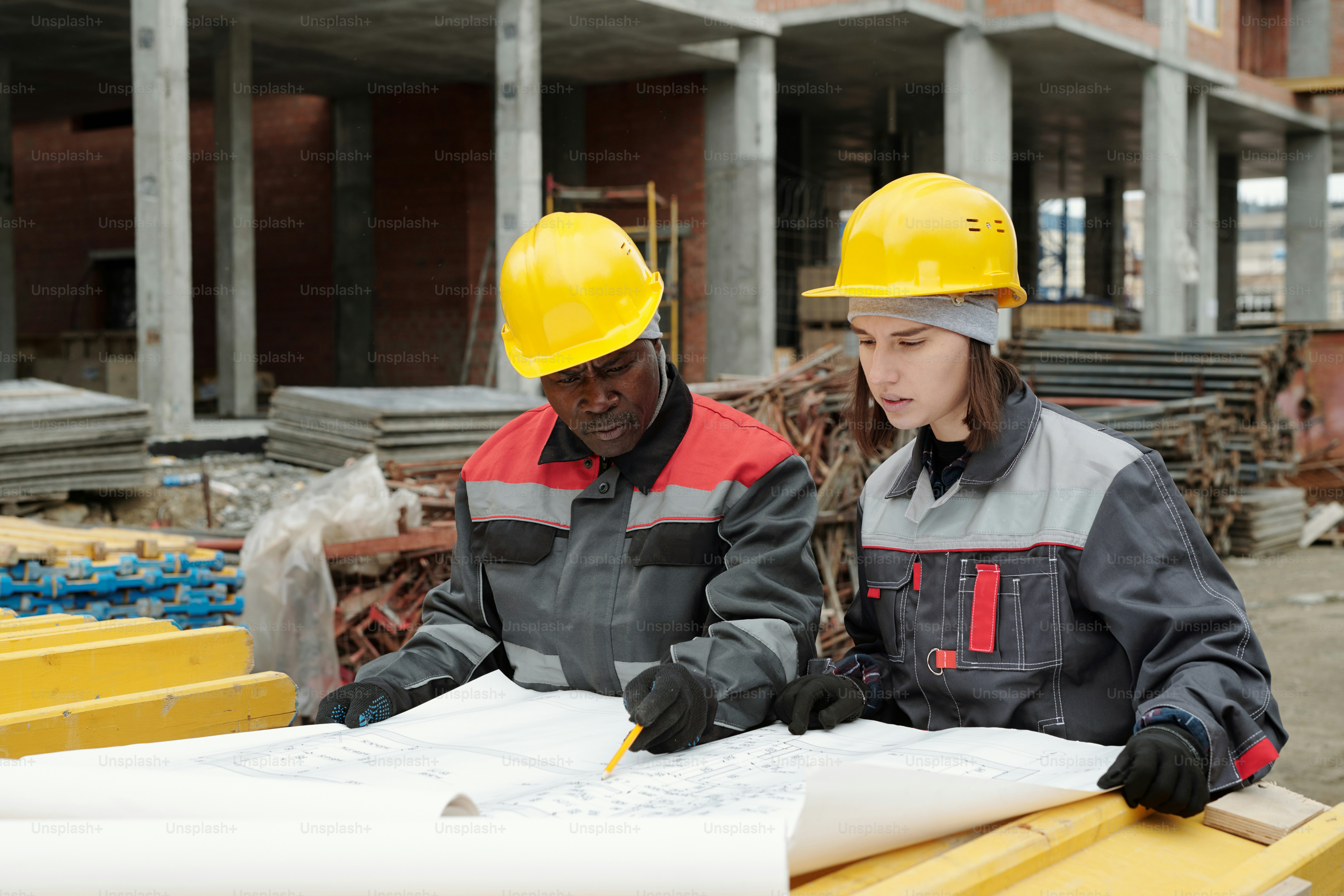 Selbstbewusster, reifer Vorarbeiter in Uniform, der jungen weiblichen Untergebenen die Skizze erklärt und gleichzeitig auf einen Bauplan mit Architekturplan zeigt