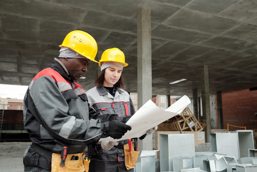 Dois engenheiros inter-raciais sérios em capacetes e roupas de trabalho olhando para o plano de construção em planta enquanto estão no canteiro de obras