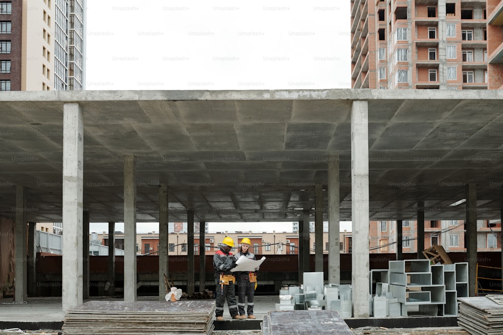 Dois construtores inter-raciais em roupas de trabalho discutindo esboço de novo edifício no canteiro de obras enquanto estão dentro de uma estrutura de concreto inacabada