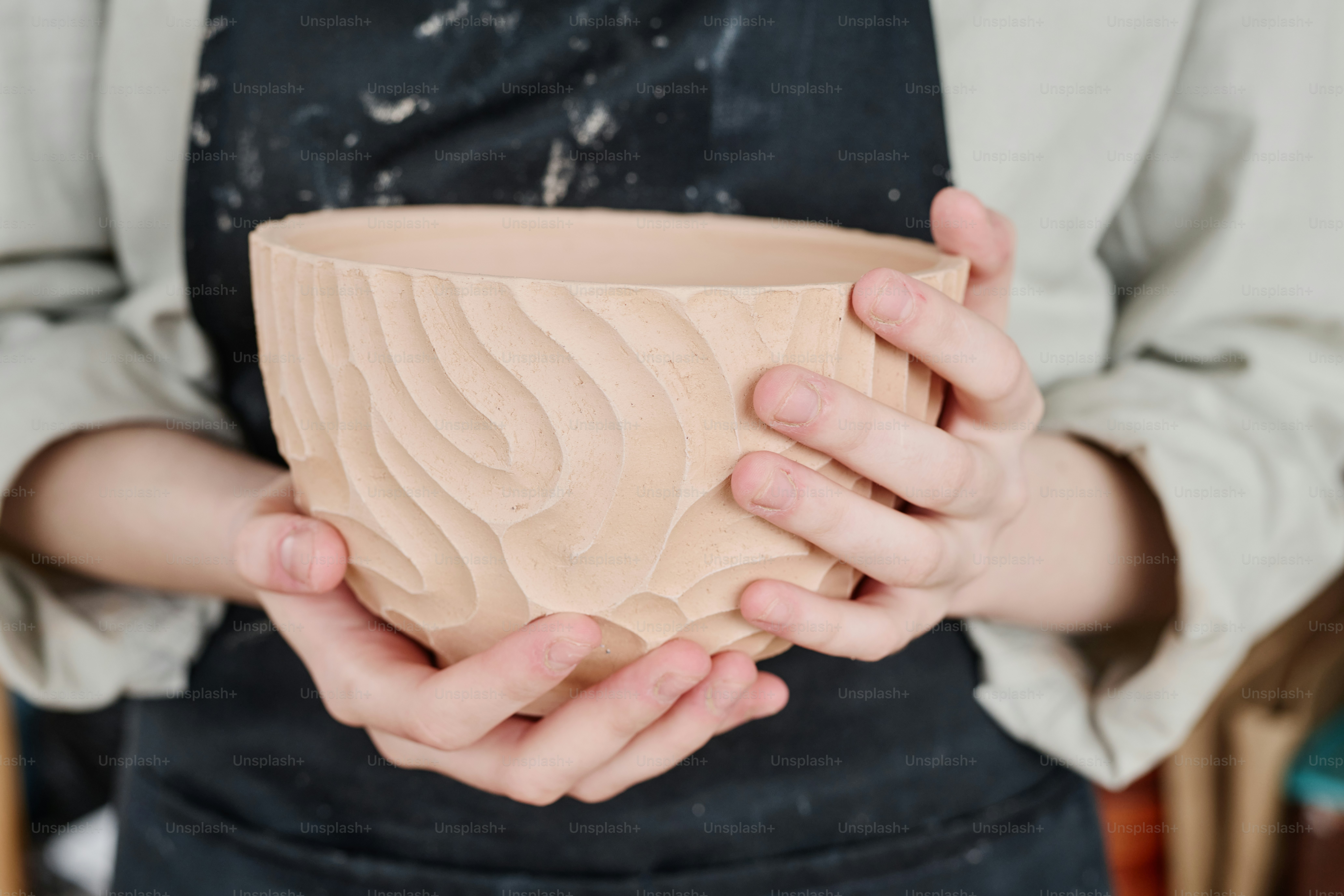 Hands of young creative female artisan or potter in workwear holding handmade clay bowl with carved ornaments on side surface