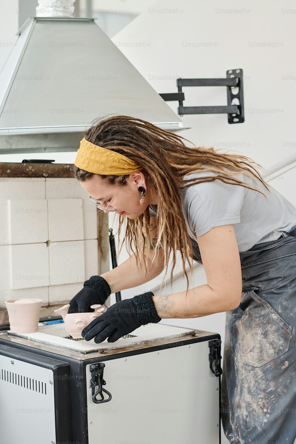 Joven mujer creativa en ropa de trabajo y guantes negros gruesos protectores inclinados sobre equipos de calefacción de barro en el taller