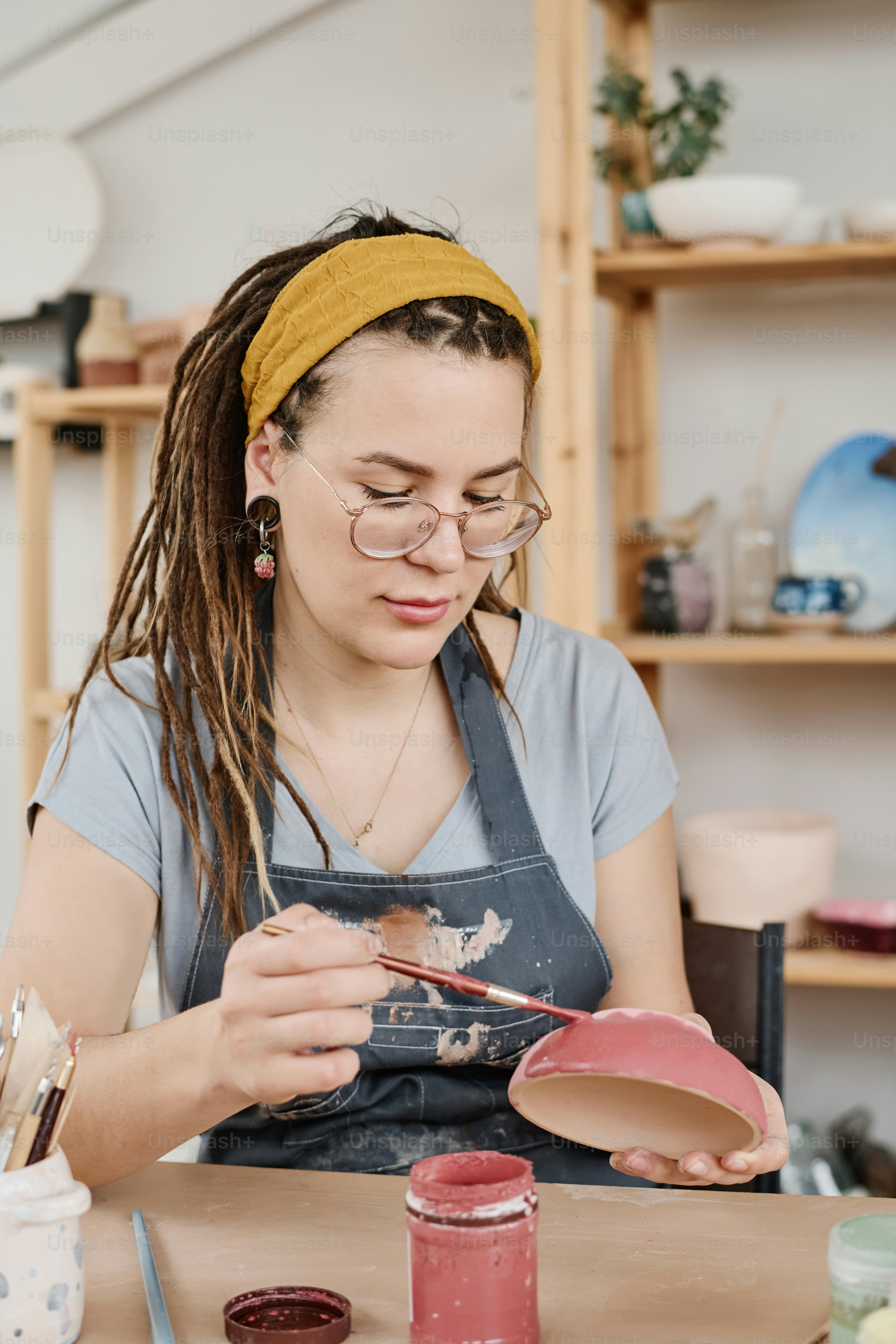 Young woman in casualwear and eyeglasses painting clay bowl with gouache or acrylic paint of marsala color by workplace