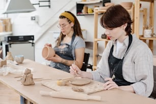 Mulher nova em workwear sentado pela mesa e ornamento de corte na peça de trabalho de argila contra colega com tigela criando novos produtos