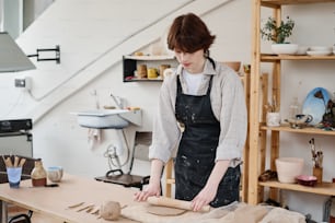 Jovem artesã ou proprietária de loja de barro no avental preto achatando pedaço de argila com alfinete enquanto está de pé ao lado da mesa