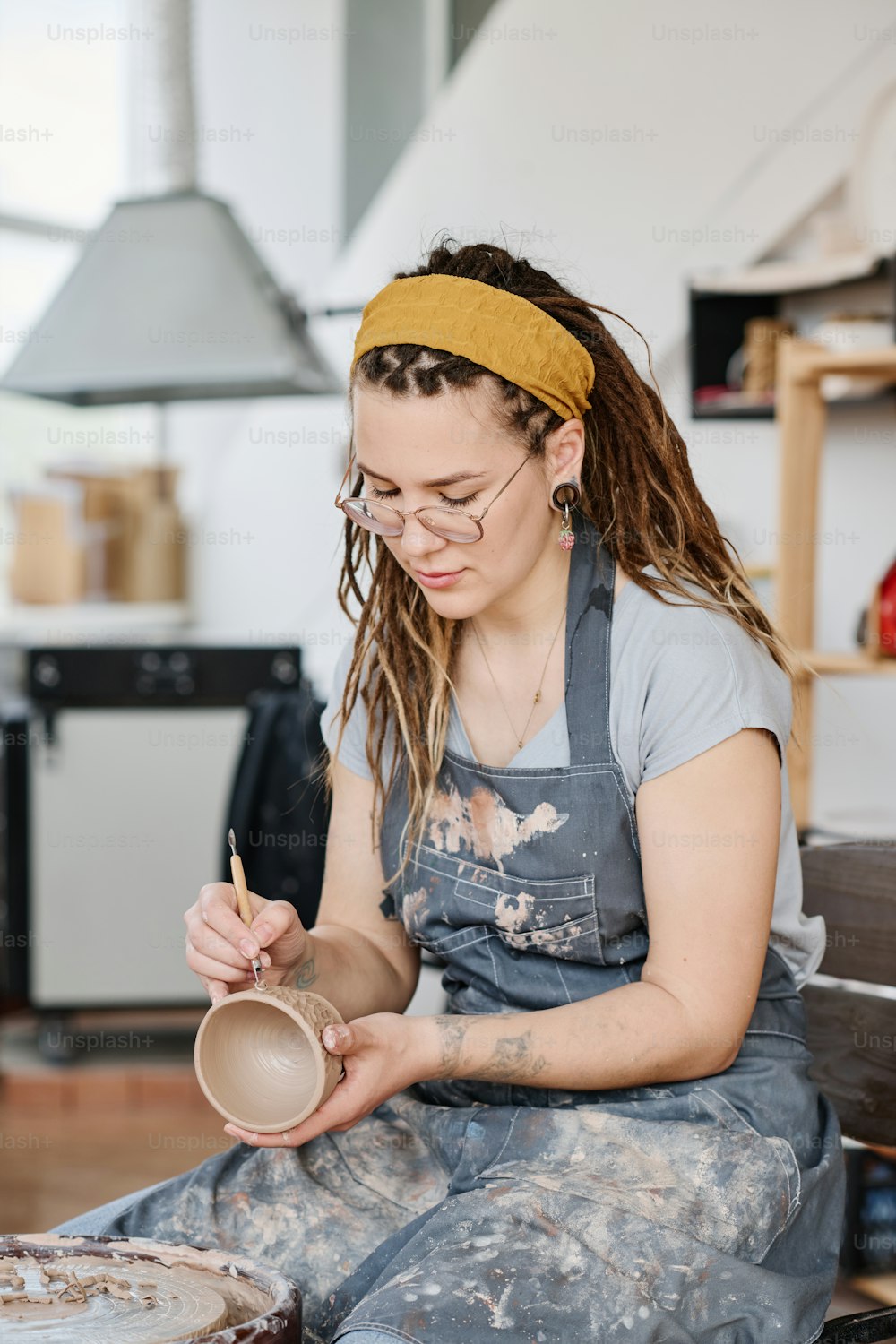 Alfarera creativa en ropa de trabajo tallando patrones decorativos en una taza de arcilla hecha a mano mientras está sentada frente a la rueda de cerámica