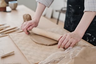 Hände einer jungen Handwerkerin mit hölzernen Nudelholz, die Ton auf Karton glätten, während sie an neuen Tonprodukten arbeiten, die in der Werkstatt verkauft werden